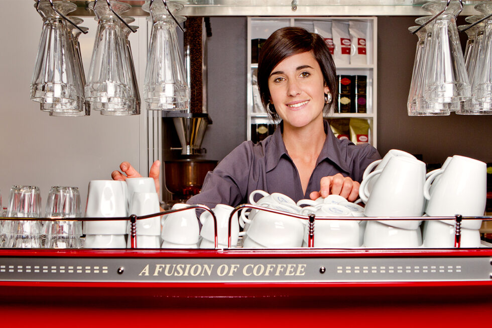 Professional commercial photograph of a smiling café barista behind an espresso machine, showcasing hospitality, customer service and workplace culture in Adelaide.