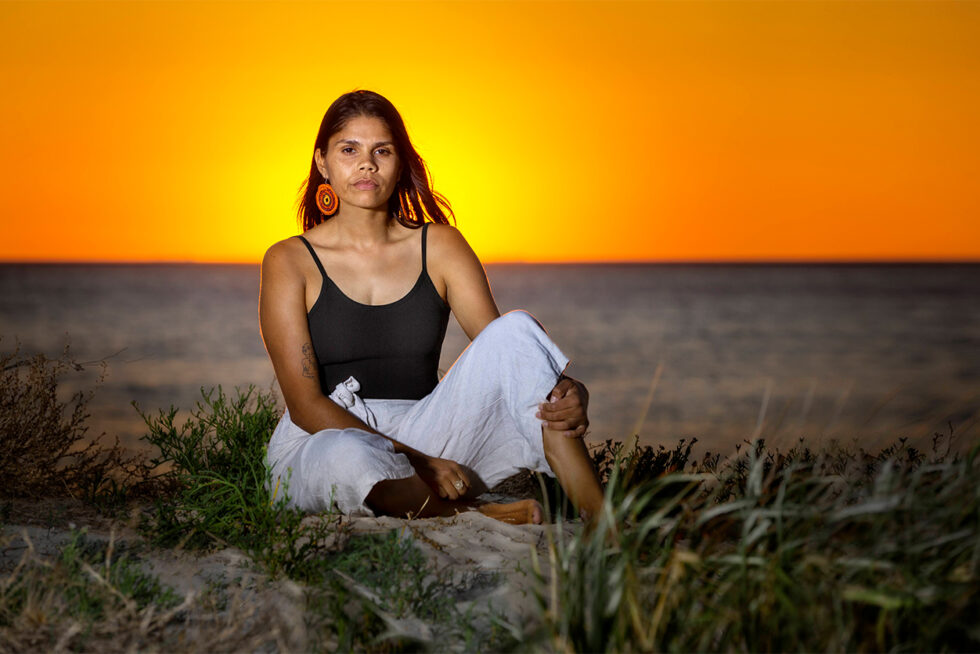 Environmental portrait of a First Nations woman seated on coastal sand dunes at sunset in South Australia.