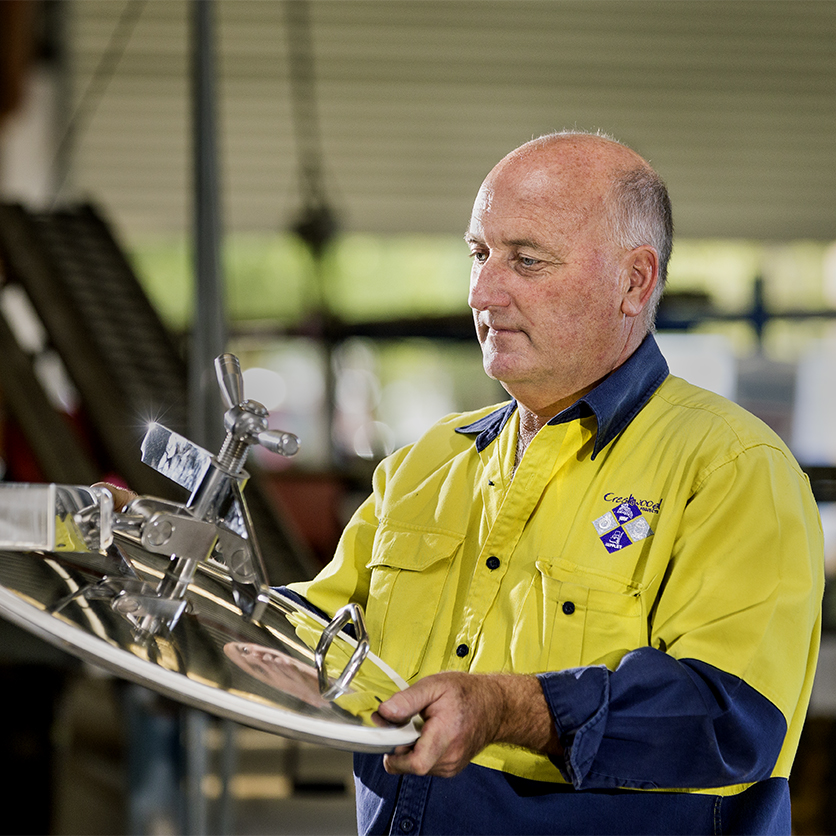 A male technician in a yellow and blue high-visibility work shirt inspecting a polished stainless steel pressure vessel hatch lid in a manufacturing workshop.